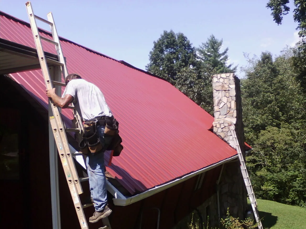 Roofer on a ladder beside a red metal roof and stone chimney — Soffit & Fascia Repair by Roofing Brothers in Coalport